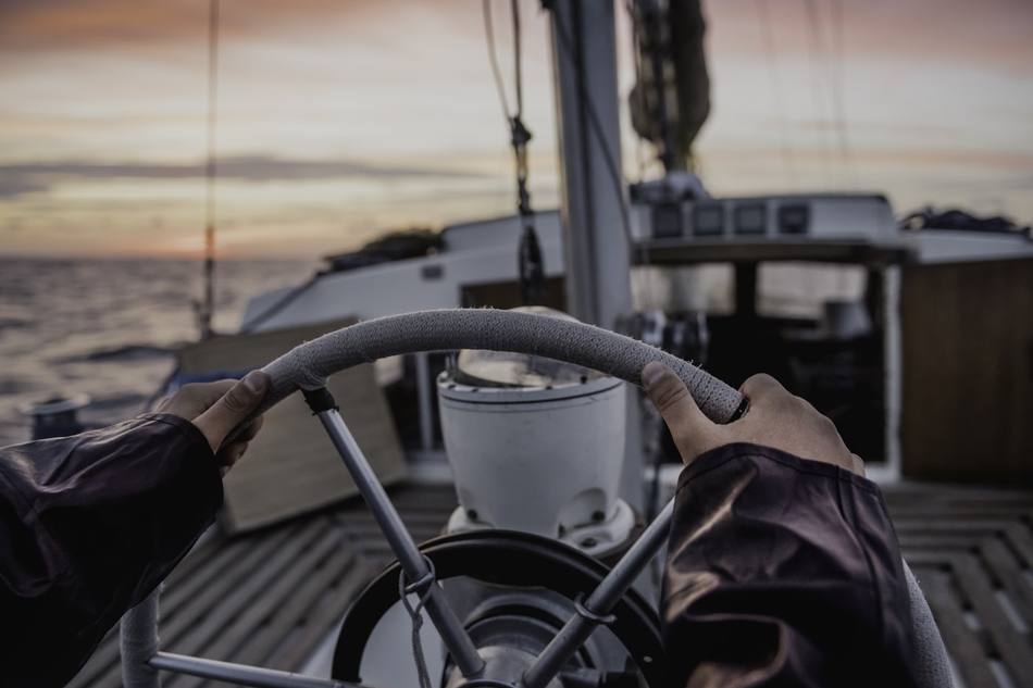 Une main tient la barre d'un bateau, la côte au loin